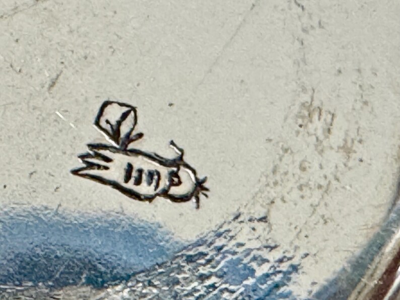 May include: Close-up of a silver-colored surface with a small, stylized bee illustration. The bee is black and has a simple, cartoonish design. The surface has a textured appearance.