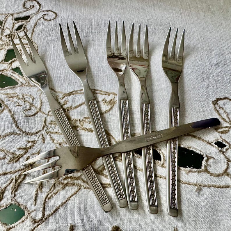 May include: A collection of six silver-toned forks, including five with a decorative geometric pattern on the handles. One fork has a different design with a dark blue handle. The forks are arranged on a white tablecloth with a floral pattern.
