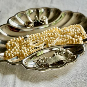 Pode incluir: Uma bandeja de metal prateado polido em forma de concha festonada. A bandeja cont&eacute;m um longo colar de p&eacute;rolas cor de creme e uma pulseira de corrente de metal prateado com textura de malha, repousando nos compartimentos curvos e reflexivos.
