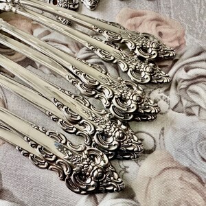 May include: Close-up of ornate silver spoons arranged diagonally. The spoons have detailed floral and scrollwork designs on the handles. The spoons are on a floral patterned fabric background.