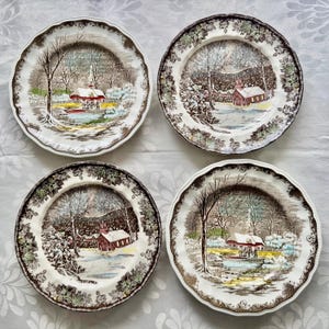 May include: Set of four vintage dinner plates featuring a winter scene with a red farmhouse, snow-covered trees, and a small pond. The plates have a brown and green floral border.