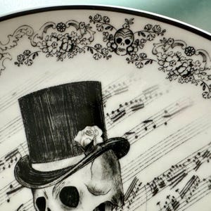 May include: A decorative plate with a black and white skull design. The skull wears a top hat with a rose, set against a backdrop of musical notes. Floral patterns and skull motifs adorn the plate's border, creating a gothic aesthetic.