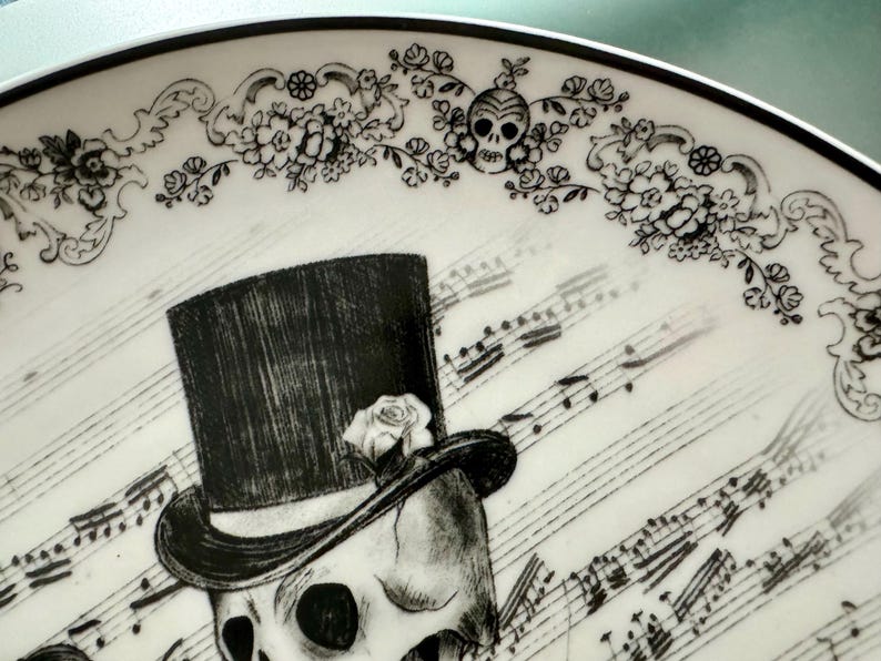 May include: A decorative plate with a black and white skull design. The skull wears a top hat with a rose, set against a backdrop of musical notes. Floral patterns and skull motifs adorn the plate's border, creating a gothic aesthetic.
