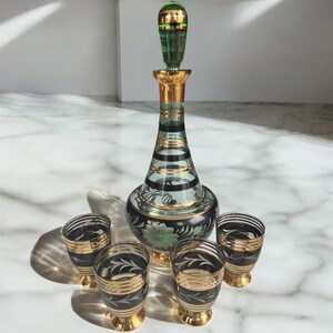 May include: A vintage glass decanter set with four shot glasses. The decanter is green and black with gold accents and a green stopper. The shot glasses are black with gold accents.