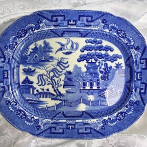 May include: An oval ceramic platter featuring a blue and white willow pattern. The design depicts a landscape with pagodas, a bridge, a willow tree, and two birds in flight. The border is decorated with a geometric pattern and floral motifs.