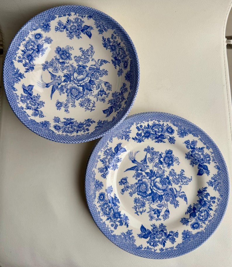 K&ouml;nnte beinhalten: Zwei blaue und wei&szlig;e Keramikteller mit einem Blumenmuster und einem Vogeldesign. Die Teller befinden sich auf einer wei&szlig;en Oberfl&auml;che.