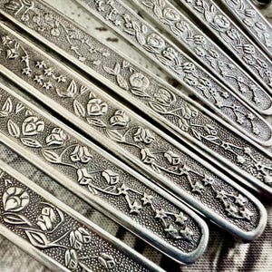 May include: A collection of silver-toned metal cutlery handles arranged in a parallel row. Each handle features a raised, textured relief pattern consisting of small five-pointed stars and blooming flower buds set against a stippled background.