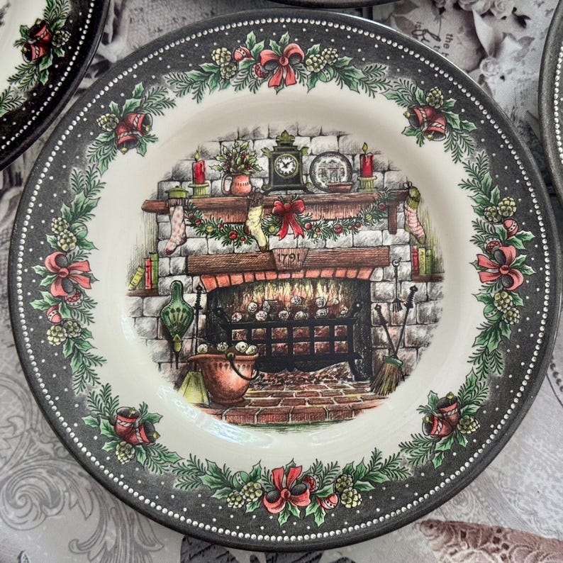 May include: A decorative plate with a detailed illustration of a fireplace scene. The plate features a black rim with white dots and a border of green leaves, red bows, and bells. The fireplace has a brick design, with a clock and candles on the mantel.