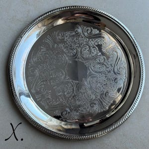 May include: A round silver-toned metal serving tray featuring a polished finish and a decorative rope-style border. The center of the tray is adorned with an intricate, embossed floral and scrollwork pattern that encircles a smooth, plain area.