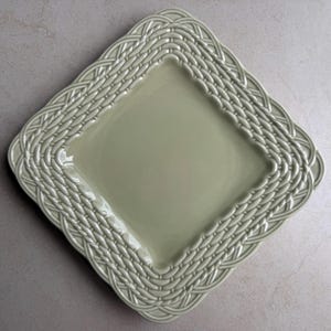 May include: A square, light sage green ceramic plate featuring a raised, embossed basket weave pattern along the entire border. The center of the plate is smooth and flat, while the edges are textured with an intricate, repeating woven design.