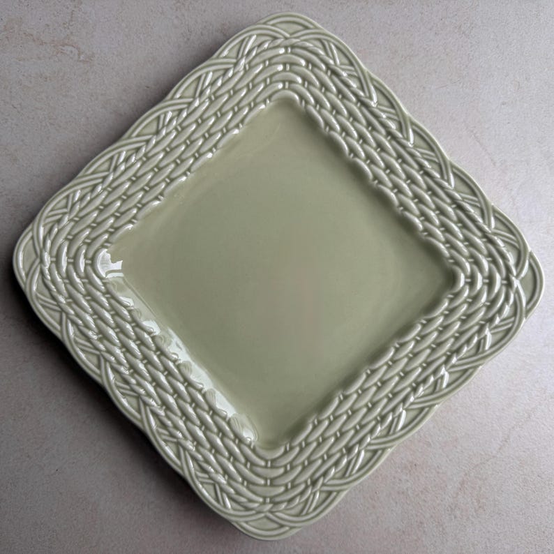 May include: A square, light sage green ceramic plate featuring a raised, embossed basket weave pattern along the entire border. The center of the plate is smooth and flat, while the edges are textured with an intricate, repeating woven design.