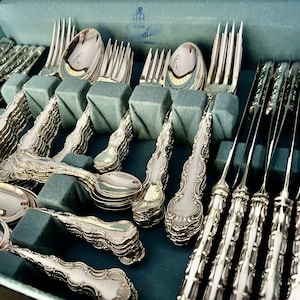 May include: A complete set of ornate silver flatware, including forks, spoons, and knives, neatly arranged in a blue velvet-lined case. The silverware features detailed, decorative handles and a polished, reflective surface, suggesting a formal dining setting.