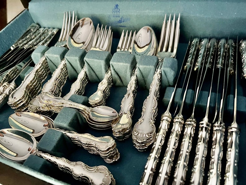 May include: A complete set of ornate silver flatware, including forks, spoons, and knives, neatly arranged in a blue velvet-lined case. The silverware features detailed, decorative handles and a polished, reflective surface, suggesting a formal dining setting.