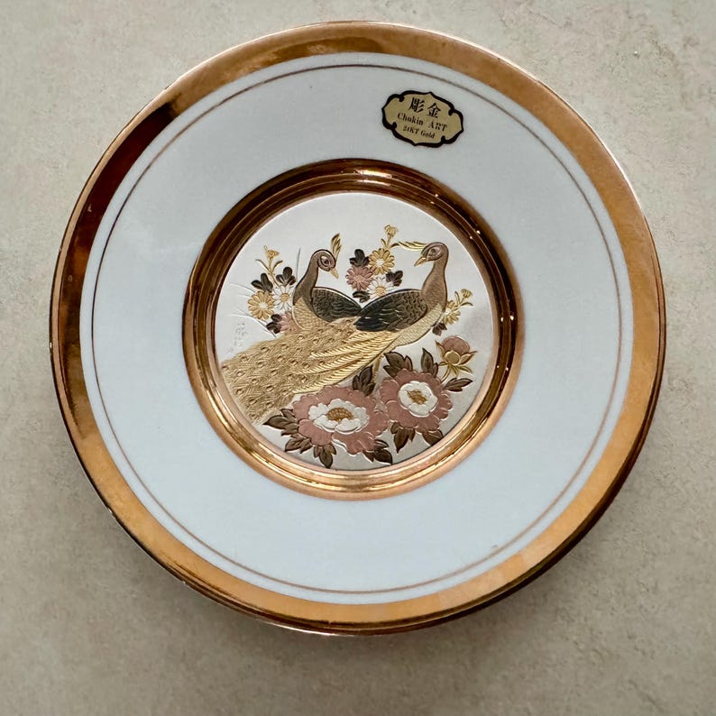 May include: A round white porcelain decorative plate featuring a central metallic silver and gold inlay of two peacocks surrounded by flowers. The plate has a wide gold rim and a black label at the top with the text Chokin ART 24KT Gold.