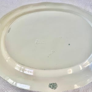 May include: An oval-shaped white ceramic serving platter with a scalloped edge. The underside features a small green circular maker's mark near the bottom rim and a faint embossed stamp in the centre of the smooth, glossy surface.