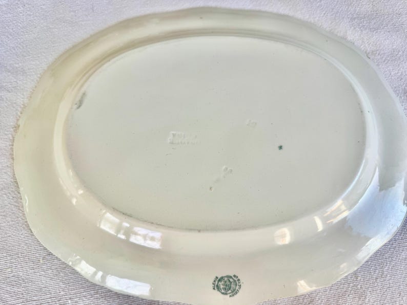 May include: An oval-shaped white ceramic serving platter with a scalloped edge. The underside features a small green circular maker's mark near the bottom rim and a faint embossed stamp in the centre of the smooth, glossy surface.
