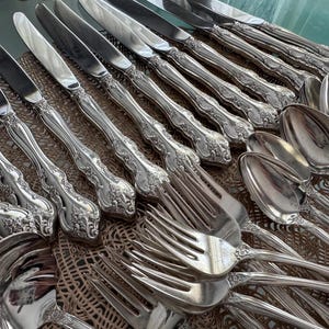May include: An assortment of antique silverware, including knives, forks, and spoons, arranged on a textured surface. The silverware has ornate, detailed handles and shiny, reflective surfaces. The collection suggests a formal dining setting.