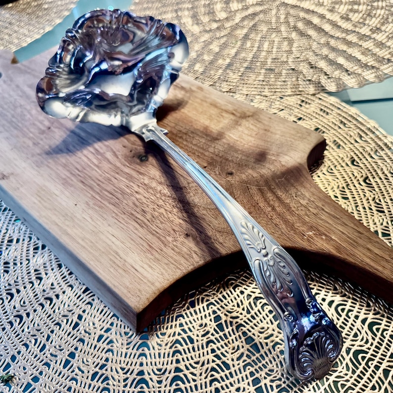 May include: A silver serving spoon with an ornate, decorative handle. The spoon has a wide, shallow bowl with a scalloped edge.