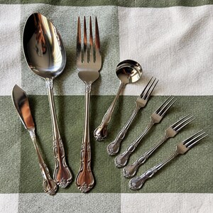 May include: A set of seven silver-toned stainless steel utensils featuring a decorative floral motif at the base of each handle. The collection includes one large spoon, one large fork, one small ladle, one butter knife, and three small forks.