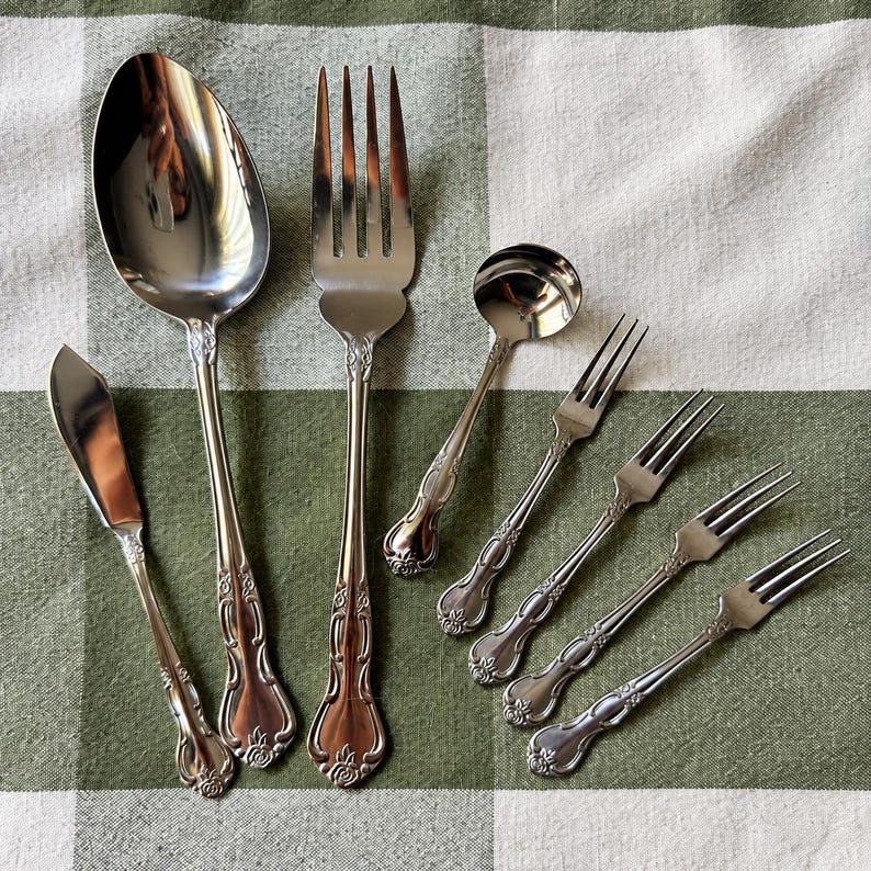May include: A set of seven silver-toned stainless steel utensils featuring a decorative floral motif at the base of each handle. The collection includes one large spoon, one large fork, one small ladle, one butter knife, and three small forks.