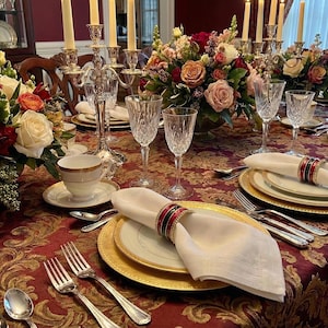 May include: A dining table set for a formal meal. The table features a burgundy and gold patterned tablecloth, gold-rimmed plates, crystal glassware, and white linen napkins. Floral centerpieces and candelabras create an elegant atmosphere for the occasion.
