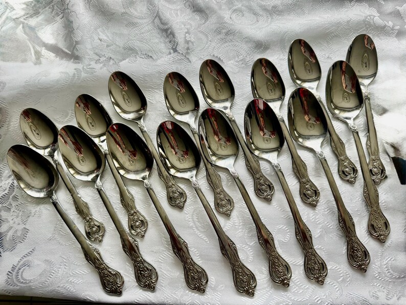 May include: A collection of silver spoons arranged on a white, patterned tablecloth. The spoons have a reflective surface and ornate, detailed handles. The spoons are likely for serving or dining.
