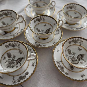 May include: Set of six white teacups and saucers with black and gold floral designs. The cups and saucers have a delicate, elegant look.
