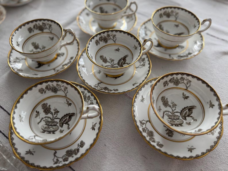 May include: Set of six white teacups and saucers with black and gold floral designs. The cups and saucers have a delicate, elegant look.