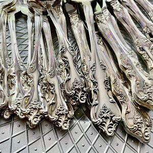 May include: Close-up of a collection of ornate silver forks. The forks have detailed floral and scrollwork patterns on the handles. The silverware is arranged on a textured, gray surface, reflecting light.
