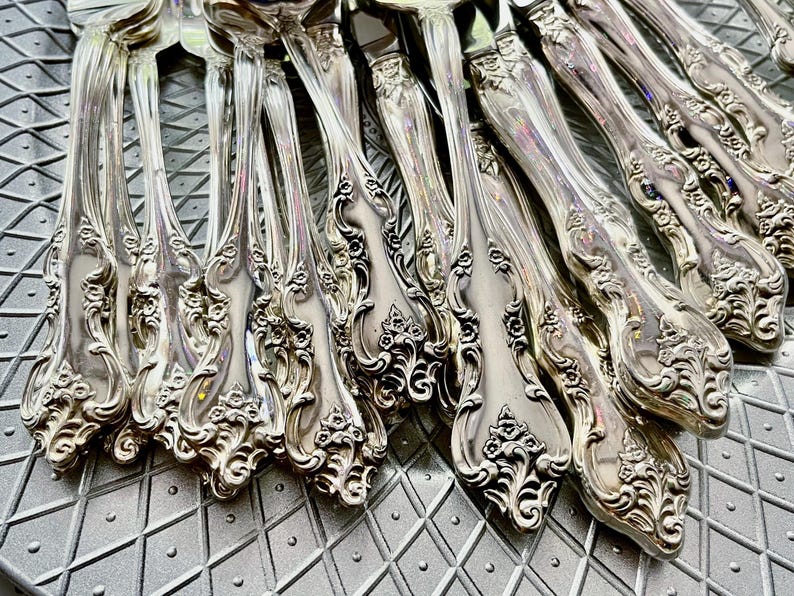 May include: Close-up of a collection of ornate silver forks. The forks have detailed floral and scrollwork patterns on the handles. The silverware is arranged on a textured, gray surface, reflecting light.
