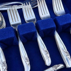 May include: A set of silver-toned metal flatware including forks and a spoon arranged in a blue velvet-lined case. Each piece features a decorative floral motif at the base of the handle and vertical grooved lines along the stem of the utensil.