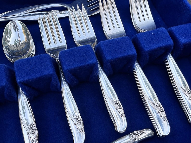 May include: A set of silver-toned metal flatware including forks and a spoon arranged in a blue velvet-lined case. Each piece features a decorative floral motif at the base of the handle and vertical grooved lines along the stem of the utensil.