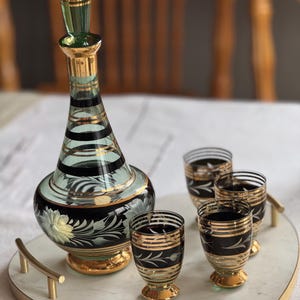 May include: A set of four clear glass shot glasses with black and gold accents on a white marble heart-shaped tray with a gold rim. The glasses are arranged in a semi-circle around a green and gold glass decanter with a black and gold floral design.