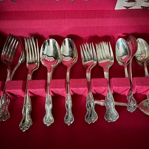May include: A set of silver-toned metal flatware including forks and spoons with ornate floral detailing on the handles, arranged in a storage case lined with vibrant red fabric. The utensils feature polished surfaces and decorative scrollwork.