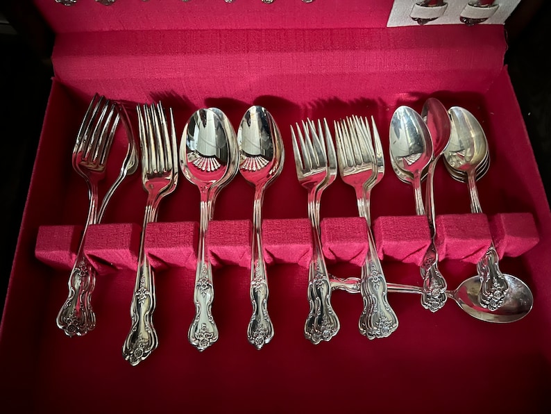 May include: A set of silver-toned metal flatware including forks and spoons with ornate floral detailing on the handles, arranged in a storage case lined with vibrant red fabric. The utensils feature polished surfaces and decorative scrollwork.