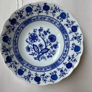 Puede incluir: Un cuenco de porcelana blanca con borde festoneado, que presenta un patr&oacute;n azul bajo vidriado. El dise&ntilde;o incluye un motivo floral central rodeado por un borde circular y un borde decorado con ilustraciones repetidas de granadas y vides.
