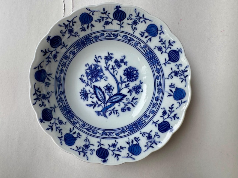 Puede incluir: Un cuenco de porcelana blanca con borde festoneado, que presenta un patr&oacute;n azul bajo vidriado. El dise&ntilde;o incluye un motivo floral central rodeado por un borde circular y un borde decorado con ilustraciones repetidas de granadas y vides.