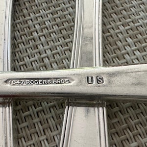 May include: Close-up of silver cutlery with the markings "1847 ROGERS BROS." and "IS" stamped on the handle. The cutlery has a classic design with a ridged pattern along the edges. The background is a woven texture.