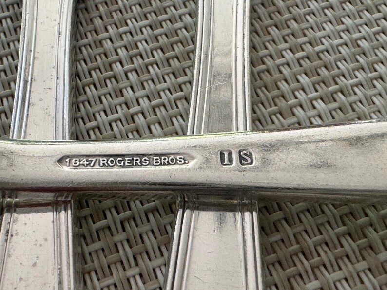May include: Close-up of silver cutlery with the markings "1847 ROGERS BROS." and "IS" stamped on the handle. The cutlery has a classic design with a ridged pattern along the edges. The background is a woven texture.