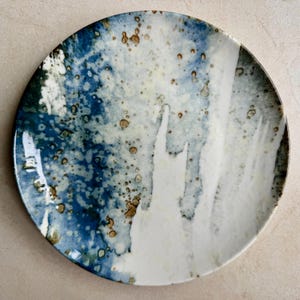 May include: A round ceramic plate featuring a mottled glaze in shades of deep blue, cream, and white. The surface displays a textured, abstract pattern with scattered brown speckles and smooth, flowing color transitions across the entire face.