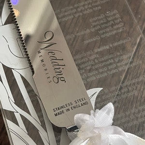 May include: A silver knife with a white handle and a white bow. The knife has the words "Wedding Memories" and "Stainless Steel Made in England" engraved on it.