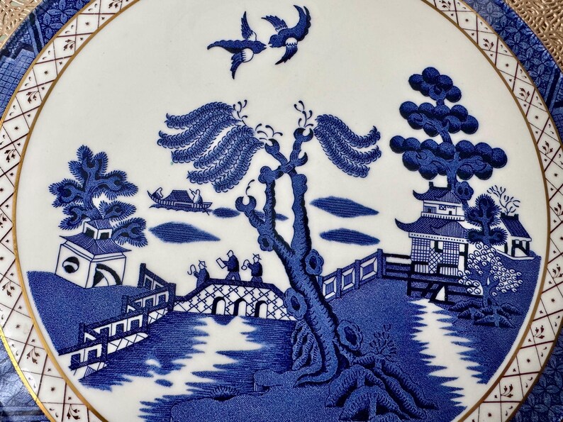 May include: Blue and white porcelain plate with a traditional willow pattern design. The pattern features a bridge, a willow tree, a pagoda, and two birds in flight.