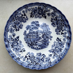 May include: A round ceramic bowl with a scalloped edge, featuring a white base and a dark blue transferware pattern. The center depicts a landscape with a house, bridge, and swans, while the rim is decorated with dense floral arrangements.