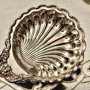 K&ouml;nnte beinhalten: Eine silberfarbene Metallschale in Form einer Jakobsmuschel. Die Schale hat eine polierte, reflektierende Oberfl&auml;che mit tiefen, strahlenf&ouml;rmigen Rillen und einen dekorativen, verzierten Griffbereich mit komplexen Schn&ouml;rkeln am Rand.