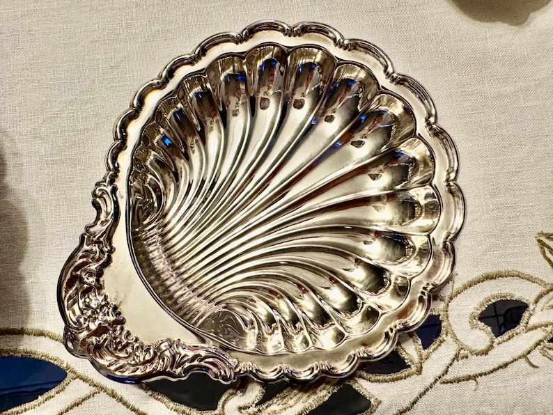 K&ouml;nnte beinhalten: Eine silberfarbene Metallschale in Form einer Jakobsmuschel. Die Schale hat eine polierte, reflektierende Oberfl&auml;che mit tiefen, strahlenf&ouml;rmigen Rillen und einen dekorativen, verzierten Griffbereich mit komplexen Schn&ouml;rkeln am Rand.