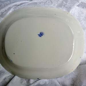 May include: An oval-shaped white ceramic platter with a smooth, glossy finish. The underside features a small, centered blue maker's mark with text and a crown emblem. The edges of the platter are slightly raised, creating a shallow basin shape.