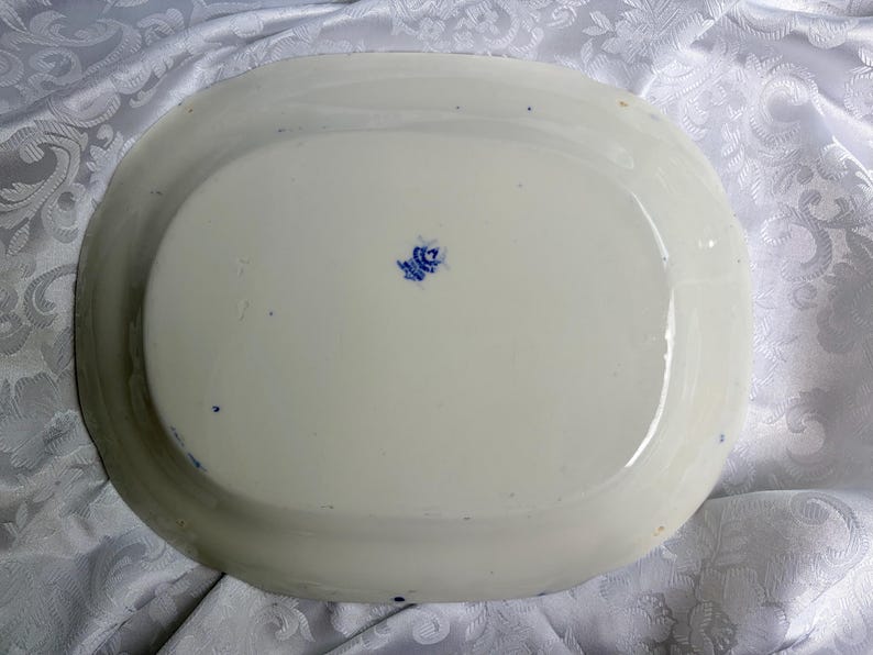 May include: An oval-shaped white ceramic platter with a smooth, glossy finish. The underside features a small, centered blue maker's mark with text and a crown emblem. The edges of the platter are slightly raised, creating a shallow basin shape.