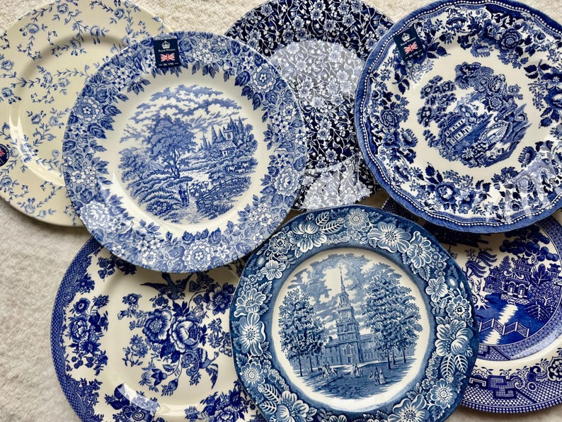 May include: A collection of six ceramic plates featuring blue and white transferware patterns. The designs include detailed landscapes, architectural scenes, and floral motifs. Each plate has a distinct decorative border and a glossy finish.