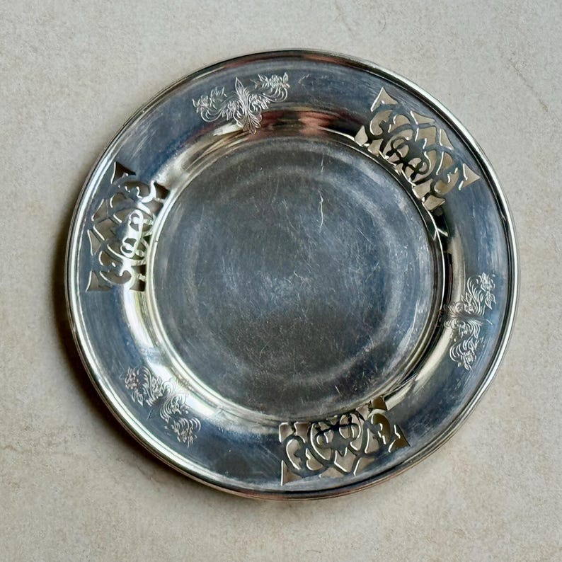May include: A round silver-toned metal plate featuring a smooth, reflective center basin and a wide, decorative rim. The rim is adorned with four symmetrical cut-out sections and intricate, embossed floral patterns between them.