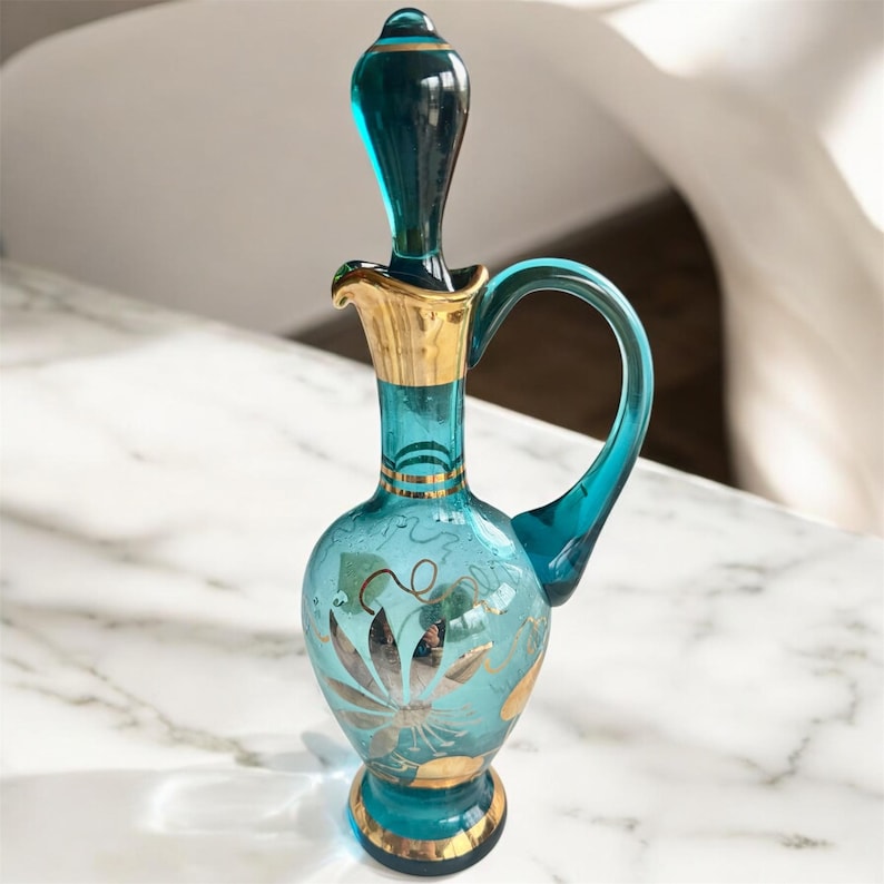 May include: A turquoise glass pitcher with a gold-trimmed spout and handle. The pitcher has a floral design in gold paint.